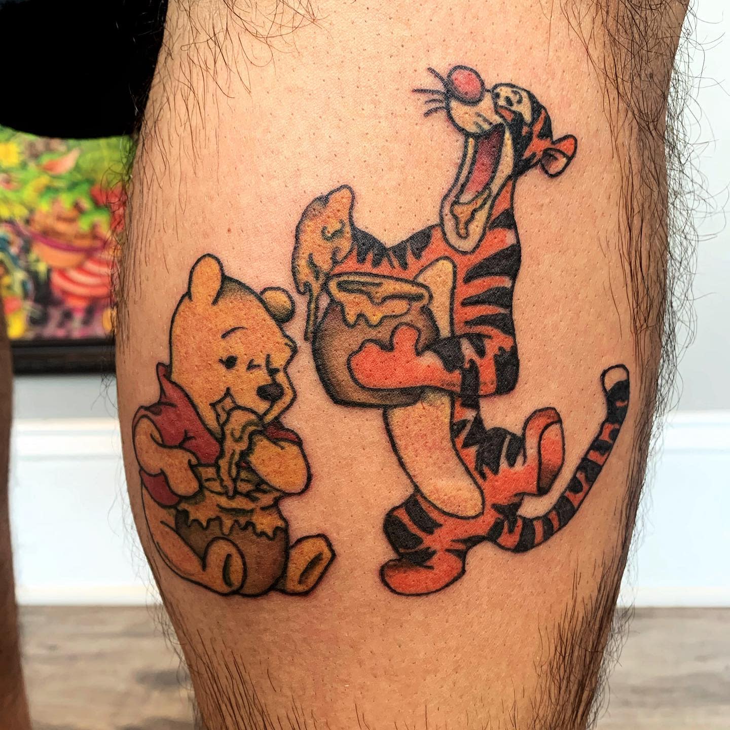 Tattoo of Winnie the Pooh and Tigger enjoying honey together on a person's leg